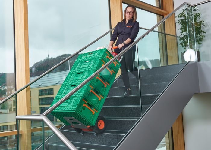 Op de foto ziet u een vrouw die een CargoMaster trapklimmer voor ladingen gebruikt om verschillende dozen met boodschappen een trap op te transporteren. U kunt ook de klimpoten van de trapklimmer zien en de veiligheidsremmen, die automatisch gestopt zijn aan de rand van de trap.