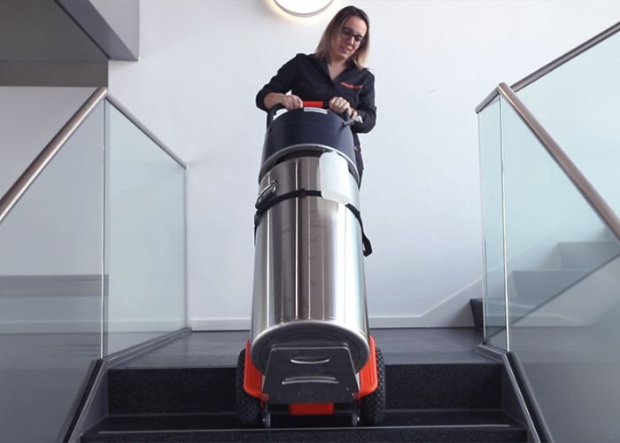 Op de foto is een vrouw te zien die een CargoMaster trapklimmer voor lasten gebruikt om een zuurstoffles een trap op te transporteren. U kunt ook de klimpoten van de trapklimmer zien en de veiligheidsremmen, die automatisch zijn gestopt aan de rand van de trap.