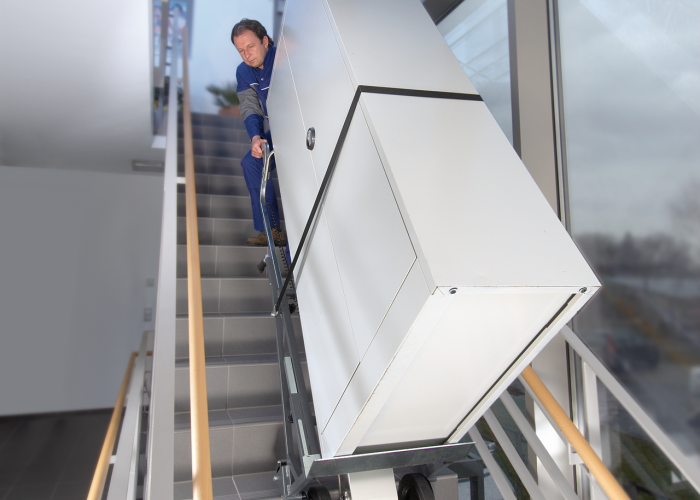 Op de foto is de CargoMaster trapklimmer in actie te zien met een grote kast die via moderne trappen in een gemeentehuis naar boven wordt getransporteerd. 