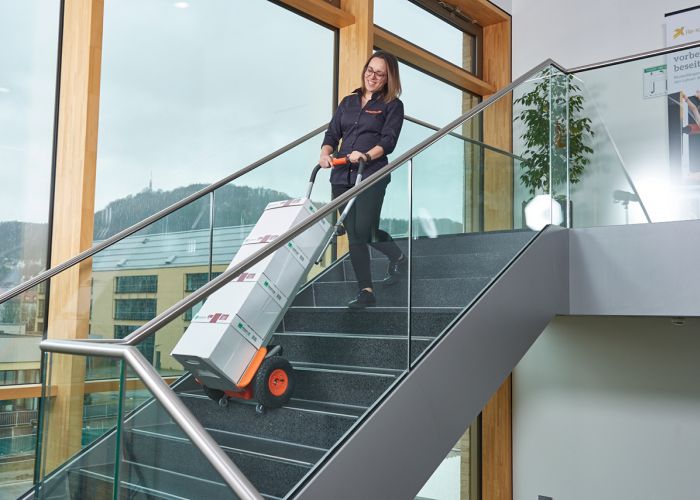 Op de foto is een vrouw te zien die een CargoMaster trapklimmer voor ladingen gebruikt om een aantal dozen kopieerpapier een trap op te transporteren. U kunt ook de klimpoten van de trapklimmer zien en de veiligheidsremmen, die automatisch gestopt zijn aan de rand van de trap.