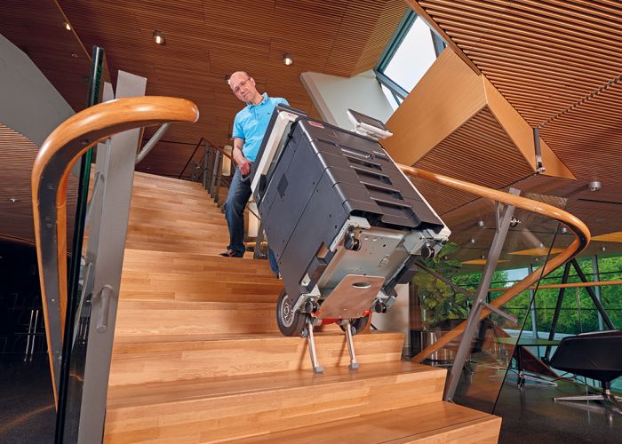 The picture shows the CargoMaster stair climber being used with a large photocopier being transported up modern wooden stairs in a town hall.